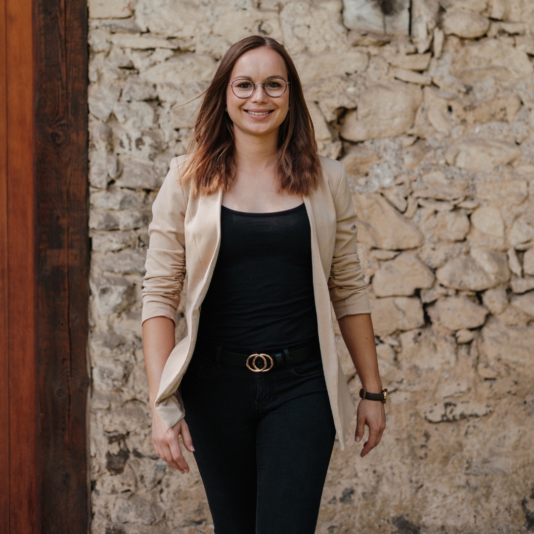 Person mit Brille steht lächelnd vor einer rustikalen Steinwand und trägt einen hellen Blazer über dunkler Kleidung.